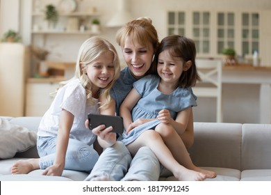 Happy Mom And Two Daughters Watching Funny Video On Smartphone Sitting On Comfortable Couch In Cozy Living Room. Smiling Mother And Children Enjoying Cartoons On Mobile Phone At Weekend