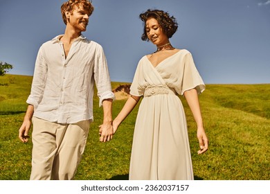 happy moment of interracial newlyweds in boho style attire holding hands in field, rural wedding - Powered by Shutterstock