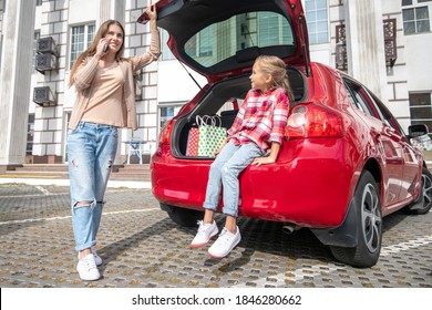 Happy Moments. Fair-haired Girl Sitting In Car Boot, Her Mom Talking On The Phone