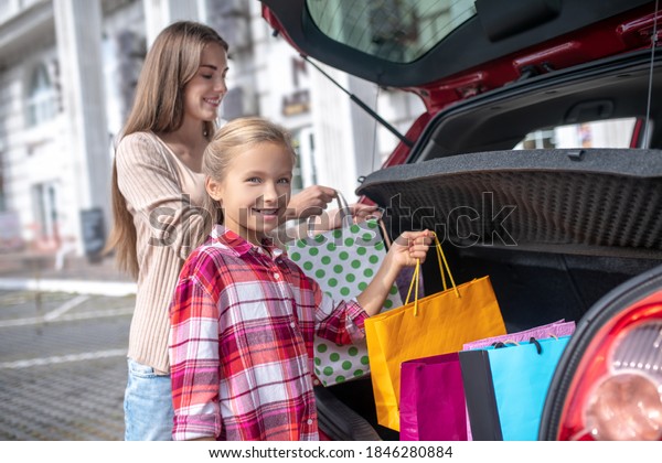 Happy moments. Fair-haired girl and young\
female taking out shopping bags from car\
boot