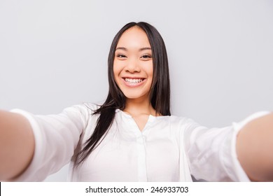 Happy moments must be saved. Cheerful young Asian woman holding camera and making selfie while standing against grey background