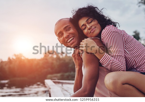 Happy moments together.\
Happy young couple embracing and smiling while sitting on the pier\
near the lake