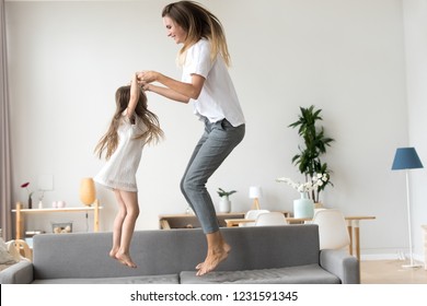 Happy Mommy And Kid Daughter Holding Hands Jumping On Sofa Together, Baby Sitter Or Mother Playing Having Fun With Cute Kid Girl At Home, Young Mum And Child Enjoy Spending Time Laughing Together