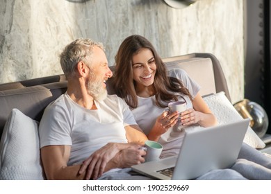 Buenos días. Alegre esposo y esposa riendo recostados con café en la cama frente al portátil en la soleada mañana