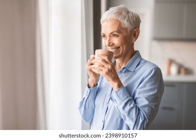 Happy morning. Relaxed smiling mature woman drinking coffee looking out window at home, enjoying domestic rest, standing with cup of steaming drink in hands, looking aside and smiling - Powered by Shutterstock