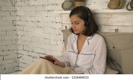 Happy morning smiling woman. Beautiful girl sitting on bed listening music with headphones singing and dancing. good-looking young lady have fun in bedroom rejoices day off at home.