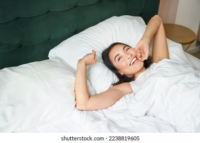 Happy mornings. Beautiful asian girl wakes up in a hotel suit, lying in white bed, stretching her arms and smiling happily. - Powered by Shutterstock