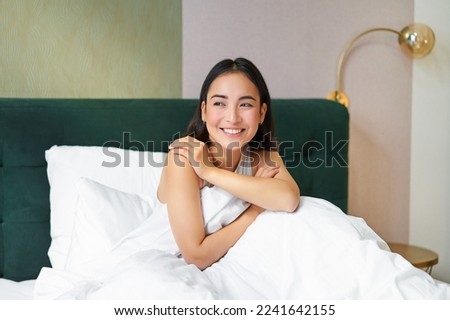 Happy mornings. Smiling asian woman wakes up in her bed, looks outside and feeld enthusiastic about her morning. Bedroom interior concept.