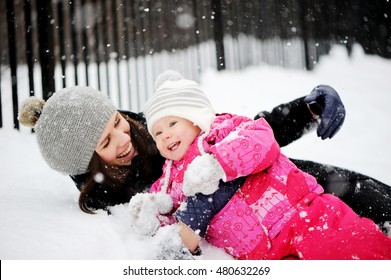 Happy Mother And Adorable Baby Girl In Cozy Winter Outfits On A Winter Walk In Snowfall.