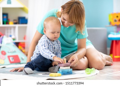 Happy Mother And Adorable Baby Playing On Floor Mat In Sunny Nursery Room. Cute Child With Carer In Creche.