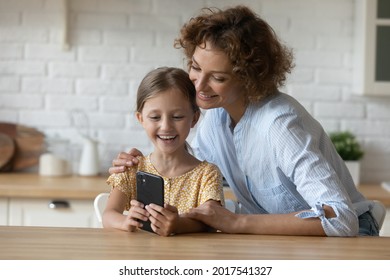 Happy Mother With Adorable Little Daughter Having Fun With Smartphone Sitting At Table In Kitchen, Smiling Mom And Preschool Kid Girl Hugging Looking At Phone Screen, Chatting Online Video Call