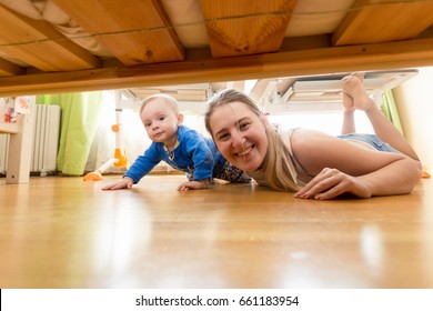 Happy Mother And Baby Boy Lying On Floor And Looking Under The Bed