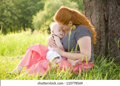 Happy mother and baby with Down syndrome hugging and kissing under the tree on the grass in the park.