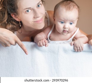 Happy Mother And Baby Lying On A White Blanket. Advertising Banner Sign - Mom Is Pointing Down And Surprised Baby Looking Down On Empty Blank Billboard Or Sign Board. Copy Space For Text And Design