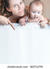 Happy Mother And Baby Lying On A White Blanket. Advertising Banner Sign - Mom Is Pointing Down And Surprised Baby Looking Down On Empty Blank Billboard Or Sign Board. Copy Space For Text And Design