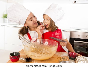 Happy Mother Baking With Little Daughter Eating Chocolate Bar Used As Ingredient While Teaching The Kid In Apron And Cook Hat And Having Fun Together