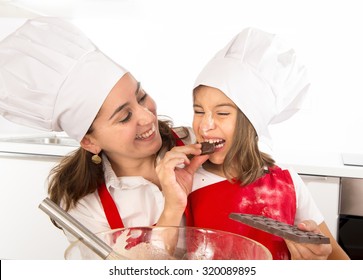 Happy Mother Baking With Little Daughter Eating Chocolate Bar Used As Ingredient While Teaching The Kid In Apron And Cook Hat And Having Fun Together