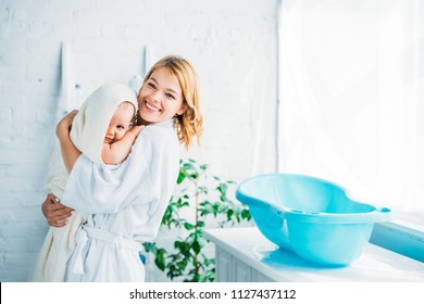 happy mother in bathrobe carrying adorable child covered in towel near plastic baby bathtub