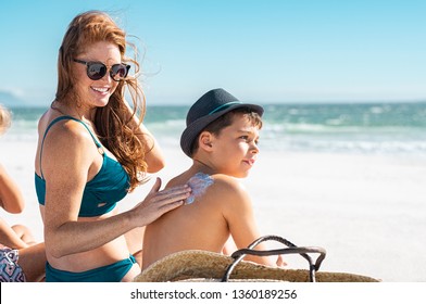 Happy mother in blue bikini sitting on beach applying sunblock cream on son's shoulder with copy space. Cute boy wearing panama hat looking away while woman applying suntan lotion on his shoulder. 