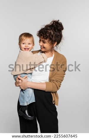 happy mother in casual clothes holding in arms cheerful baby girl in sweater isolated on grey