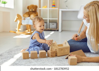 Happy Mother And Child Playing Game With Wood Cubes At Home Together. Nanny Looking After Little Kid Busy With Blocks And Toy House. Cute Toddler And Babysitter On Warm Floor In Modern Nursery Room