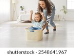 Happy mother and child playing together. Young mom pushing a plastic laundry basket with her cute little smiling cheerful kid daughter sitting inside. Family, leisure, fun concept