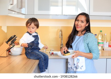 Happy Mother Cleaning Dishes At Kitchen Home With Her Kid. Mixed Race Asian-German Family Woman And Child With Housework.