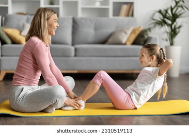 Happy mother and cute little daughter exercising at home together, sporty family mom and female child doing sit-ups, training on fitness mat in living room interior, mommy supporting kid, side view - Powered by Shutterstock