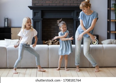 Happy mother dancing, having fun with two daughter in modern living room. Family playing funny game. Young mom and adorable cute girls moving to favorite music, enjoying weekend at home.