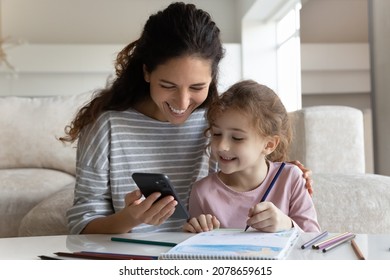 Happy Mother With Daughter Distracted From Drawing With Colorful Pencils Using Smartphone, Enjoying Leisure Time With Gadget, Smiling Mom And Adorable Girl Kid Having Fun Online Or Watching Tutorial