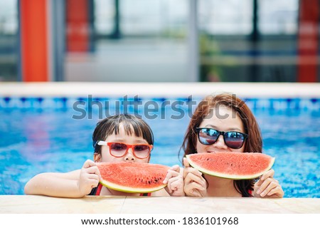 Happy mother and daughter eating watermelon after swimming in pool of spa hotel