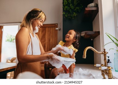 Happy mother and daughter having fun with soap bubbles in the kitchen. Cheerful mom laughing with her daughter while washing the dishes. Loving single mother bonding with her daughter. - Powered by Shutterstock