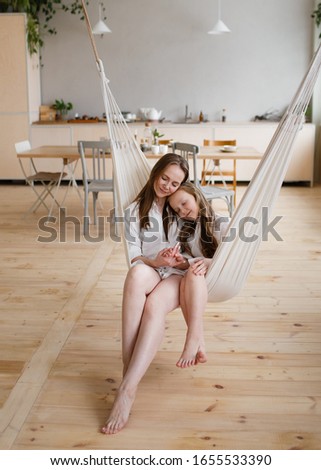 Happy mother and daughter in home white shirts relax in a hammock, have Breakfast and drink tea in the kitchen, have fun, communicate, hug in a cozy bright house, comfort, love, care, family, nature.