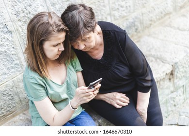 Happy Mother And Daughter Looking At A Mobile Phone Outside - Young Female Teaching Senior Woman How To Use Modern Technology - Concept Image For Elderly People Using Gadgets