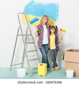 Happy Mother And Daughter Makes Repairs At Home. Smiling Woman And Girl Painting Big Ukrainian Flag On Wall At Room.