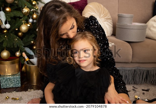 Happy mother and daughter\
sitting near Christmas tree at home. Merry Christmas and Happy\
Holidays.
