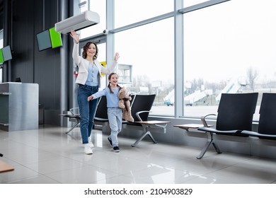 Happy Mother And Daughter Waving Hands While Walking In Airport Lounge Hall