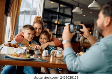 Happy mother embracing her kids while father is photographing them during family breakfast in hotel restaurant.  - Powered by Shutterstock