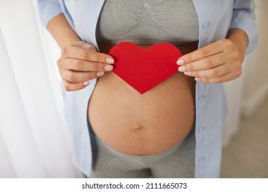 Happy Mother Expecting A Baby. Young Pregnant Woman With A Cute Bare Belly Button Holding A Red Paper Heart In Her Hands. Cropped Shot, Close Up. Pregnancy, Motherhood, Love, Care, Health Concepts