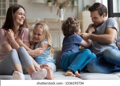 Happy mother and father having fun with little children at home, laughing parents tickling cute daughter and son, enjoying weekend with kids indoors, cuddling, playing funny game on couch