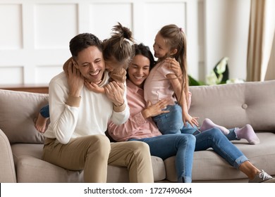 Happy mother and father hugging and cuddling with two little daughters, sitting on cozy sofa at home, smiling parents and preschool children having fun together, family enjoying free time