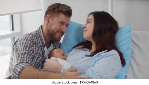 Happy Mother And Father With Newborn Baby At Hospital. Portrait Of Young Couple Holding Newborn Son Or Daughter Sitting In Hospital Bed. Parenthood And Healthcare Concept