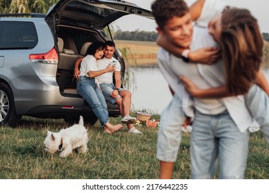 Happy Mother and Father Sitting in Trunk of Minivan Car While Children Playing with by Lake, Four Members Family and Pet Dog Having Weekend Picnic Outdoors