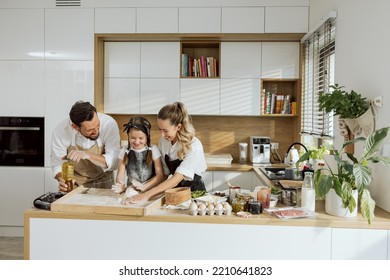 Happy mother and father teaching daughter in aprons kneading dough on wooden surface. Happy family baking cooking preparing homemade dough in modern light kitchen. - Powered by Shutterstock