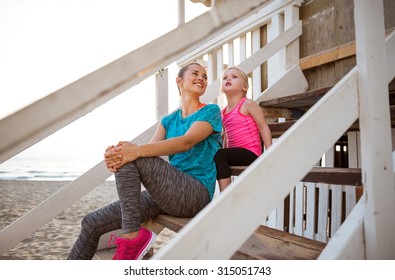 A Happy Mother In Fitness Gear Is Sitting With Her Daughter On The Wooden Steps Of Their Beach-front House. Both Are Wearing Fitness Gear. The Mother Is Looking Over Her Shoulder At Her Daughter.