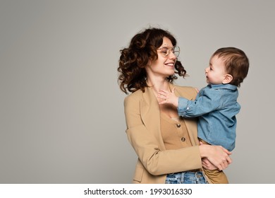 Happy Mother In Glasses And Blazer Holding In Arms Baby Boy Isolated On Grey