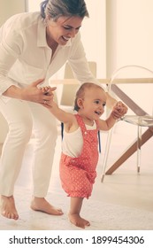 Happy Mother Helping Cute Baby Doing First Step. Lovely Barefoot Infant Learning Walking On Floor At Home And Smiling. Caring Mom Holding Daughter Hands. Family Time And Childhood Concept