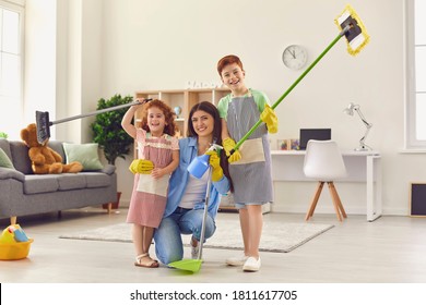 Happy Mother And Her 6 And 10 Year Old Helpers In Rubber Gloves With Floor Mops And Dustpan After Tidying Up Their House. Portrait Of Mom And Kids Smiling And Looking At Camera In Clean Living Room