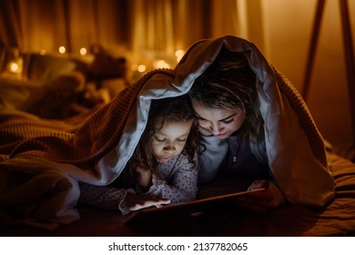 Happy mother with her little daughter lying under blanket and watching movie on tablet at home. - Powered by Shutterstock