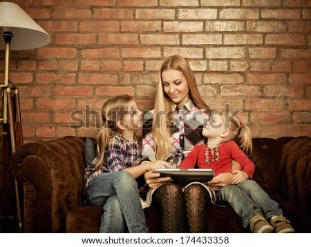 Happy mother and her little daughters sitting on a sofa at home having fun using a tablet computer. Brick background